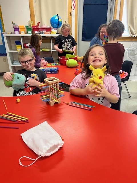 Two young children, a boy and a girl, are sitting at a red table in a classroom, smiling at the camera. The girl is holding a yellow Pikachu plush toy, and the boy is holding a blue turtle or tortoise plush toy. A wooden game with colored sticks is in the center of the table, surrounded by colored pencils.