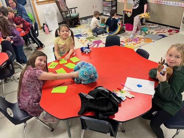 Three young girls are sitting at a large red hexagonal table in a classroom, engaged in an activity. The girl on the left is holding a large blue plush caterpillar/monster toy. The girl on the right is holding a small reindeer plush toy. Colored markers, sticky notes, and a paper with a blue circle design are on the table. Other children and toys are visible in the background.
