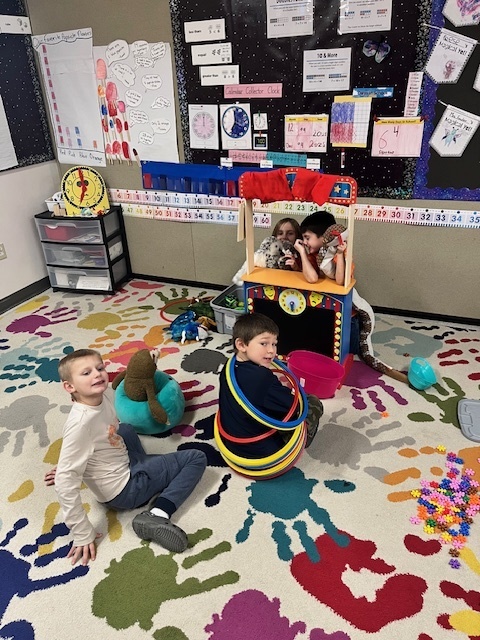 Three young boys are playing in a classroom. One boy is sitting with a plush seal or sea creature next to him. Another boy is sitting inside a stack of colorful hula hoops. A third boy and an adult are playing inside a small, colorful play theater/puppet show structure in the background. Colorful floor tiles and math charts are visible.