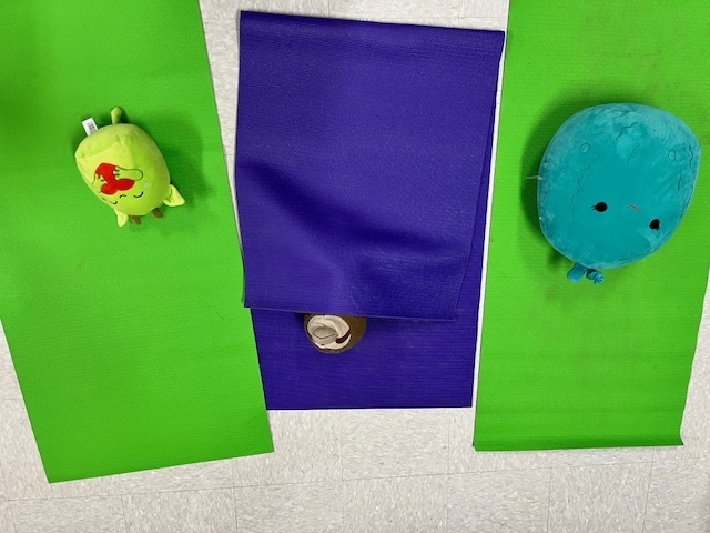 An overhead view of two green yogavocado-sa mats and one purple folded mat on a light-colored floor. A green shaped plush toy is resting on the left mat, and a blue, round-shaped plush toy is on the right mat. A small brown coconut toy is tucked under the purple mat.