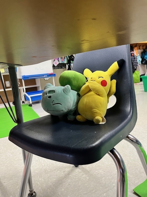 A close-up of a blue classroom chair with a green Bulbasaur plush toy and a yellow Pikachu plush toy sitting on the seat, tucked under a desk.