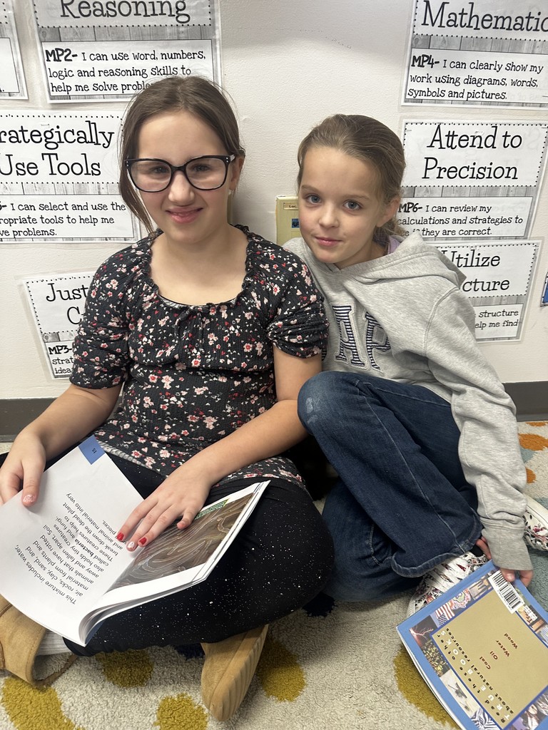 Two girls are sitting together on the classroom floor, leaning against a wall with academic posters. The girl on the left wears glasses and a dark floral top, and the girl on the right wears a gray 'A E' hoodie. They are holding an open book together.