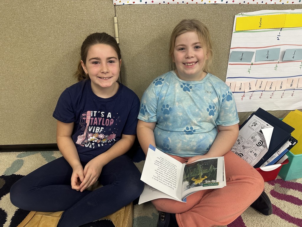 Two girls are sitting side-by-side on the classroom floor, smiling at the camera. The girl on the left wears a navy "IT'S A TAYLOR VIBE" shirt, and the girl on the right wears a light blue tie-dye shirt with paw prints. They have an open book between them.