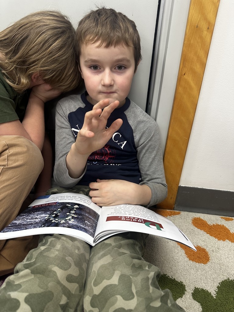 Two young boys are sitting on a colorful, patterned rug, closely reading from the same open book. They are leaning against a bulletin board with classroom rules written on it.