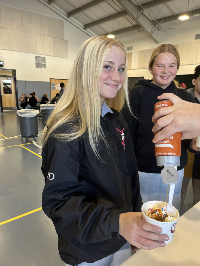 A student smiles while a staff member drizzles caramel sauce over her ice cream in a busy school gathering area.