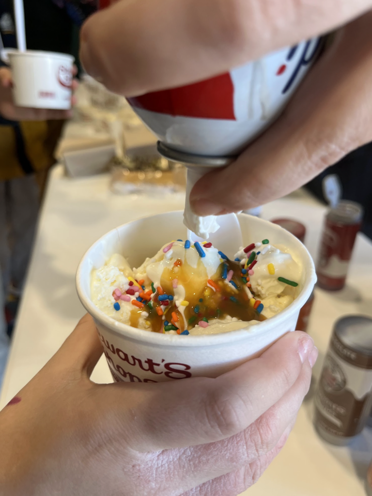 A close-up of a hand topping a cup of ice cream with whipped cream, caramel sauce, and rainbow sprinkles.