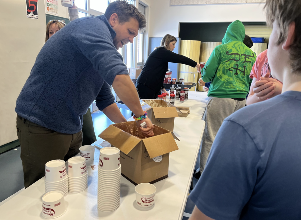A staff member scoops chocolate ice cream from a large box while students line up at a toppings table in a school cafeteria space.