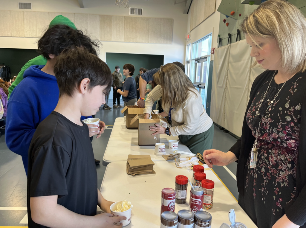 A student stands at a table holding a cup of ice cream while staff members scoop ice cream and prepare toppings for students in a school gym.