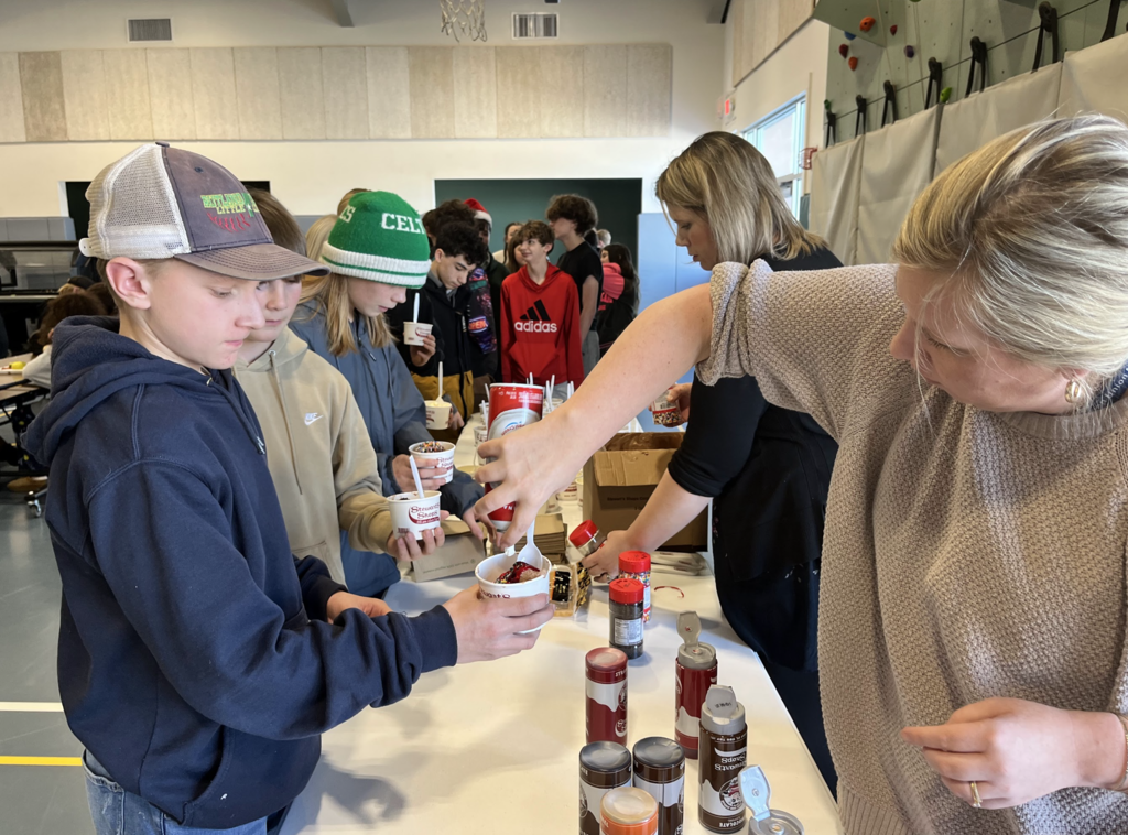 Students hold ice cream cups while a staff member adds whipped cream and toppings; a long line of students waits behind them.