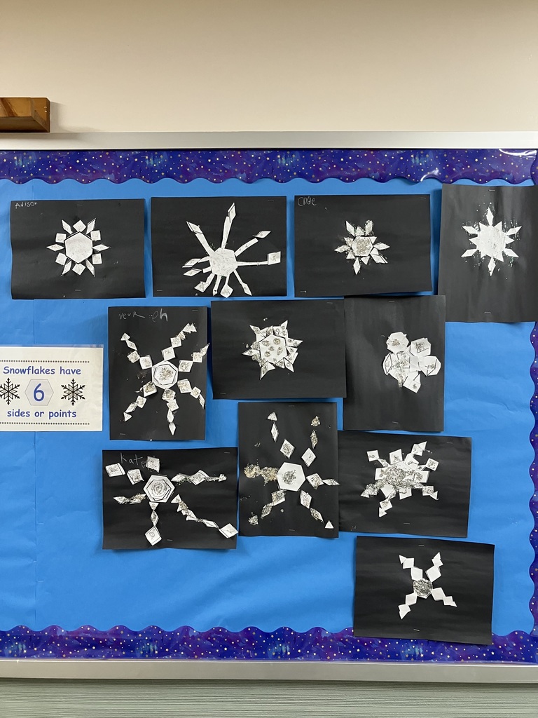 11 different student created snowflakes on black paper, they are silver, they are posted on a blue background on a bulletin board.