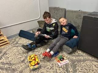 Older student sitting on cushions reading to a younger child beside a bin of toys.