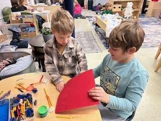 Older student helping a younger child with a red paper craft at a table.
