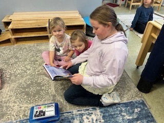 Older student sitting on a rug reading a book to two young children leaning in.