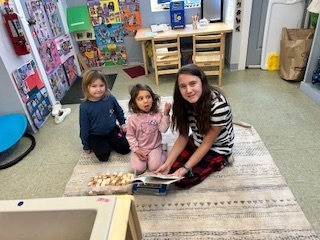 Older student on a rug reading to two younger children who sit beside her.