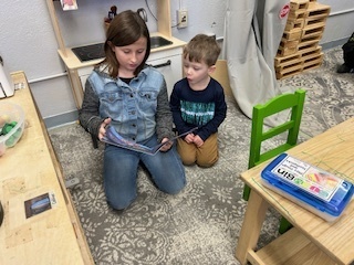 Older student kneeling and reading to a younger child standing beside her.