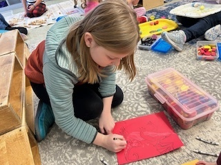 Student drawing on red construction paper while sitting on a rug with art supplies nearby.