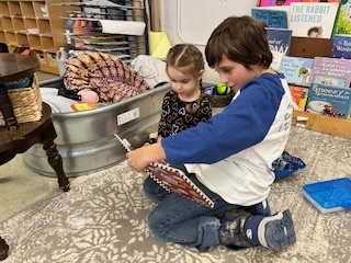 Older student kneeling on a rug reading to a younger child who watches the book.