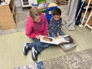Older student sitting on the floor reading a large picture book with a younger child.