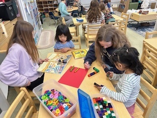 Two older students at a table helping two younger children with blocks and art materials.