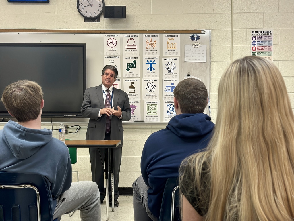 Senator Collamore speaks to students seated in a classroom, standing near a screen and a wall of career-cluster posters.