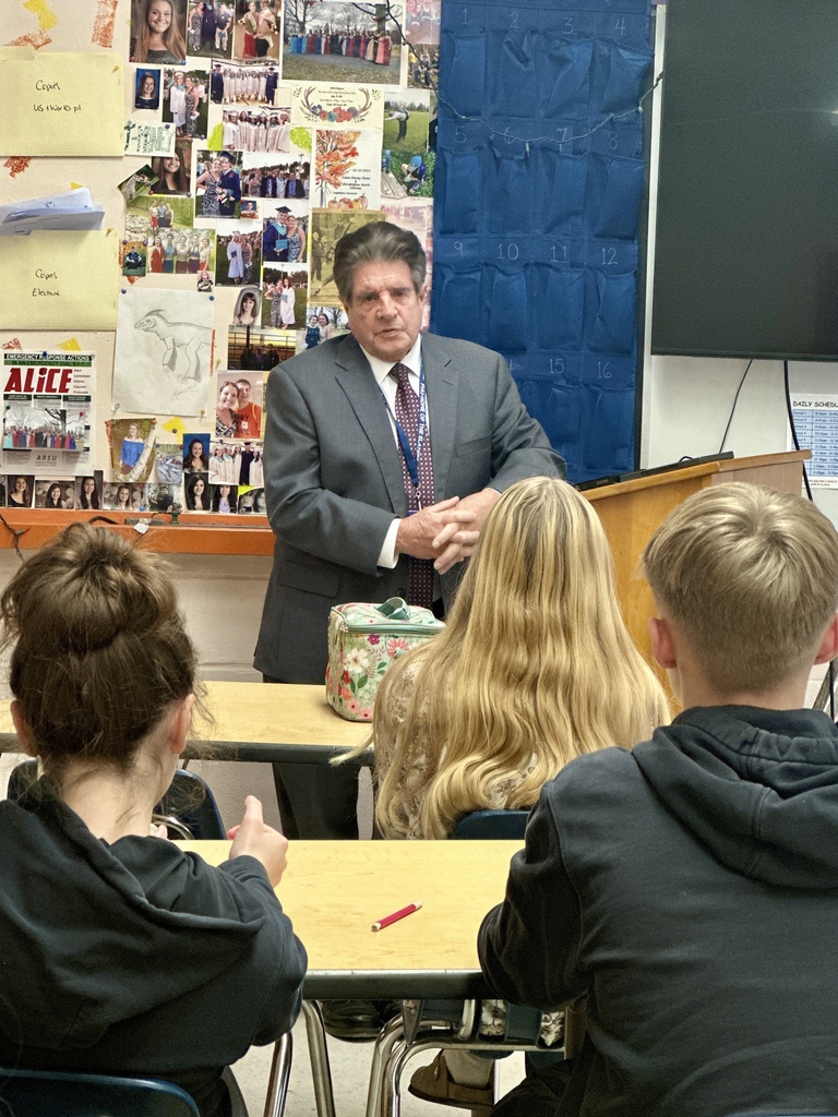Senator Collamore speaks at a podium in a classroom while students listen; a collage of photos and posters is visible on the wall behind him.