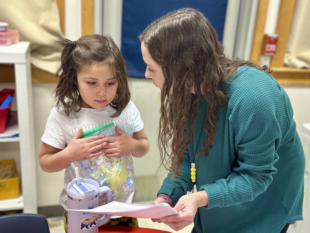 A teacher in a teal sweater bends down to review a paper with a young girl holding a plastic bag of materials.