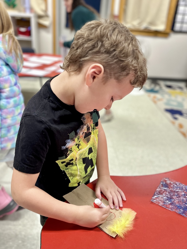 A young boy draws on a brown paper bag puppet, adding feathers at a red table.