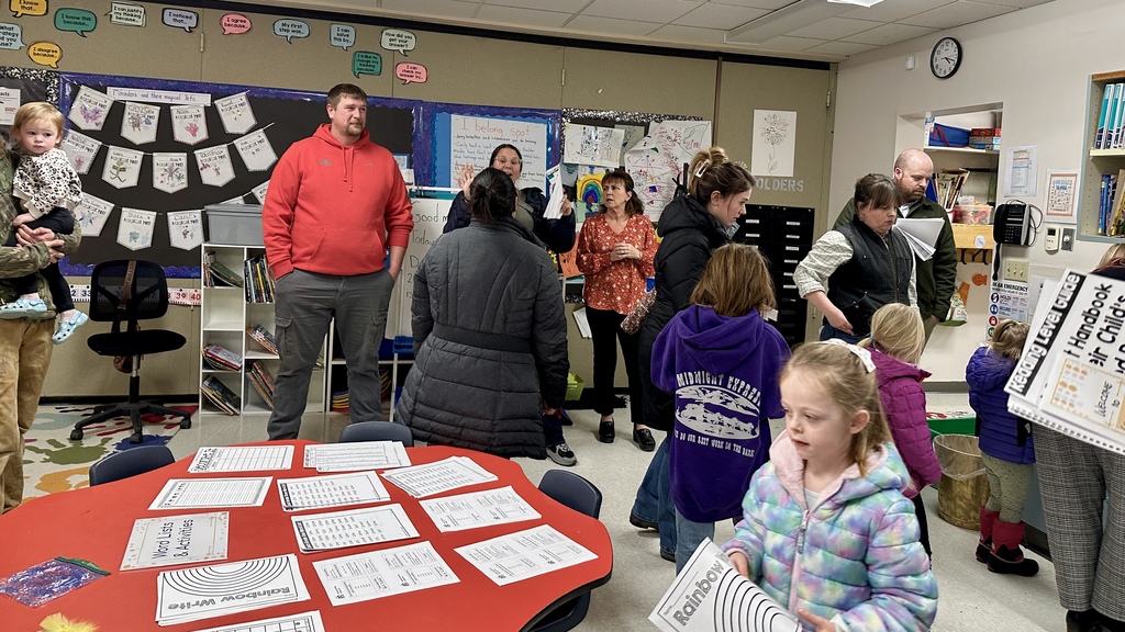 Families and students stand and talk around the classroom during an open house event with worksheets displayed on tables.