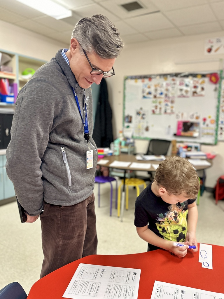 A male teacher watches a young student draw on a strip of paper at a red table.