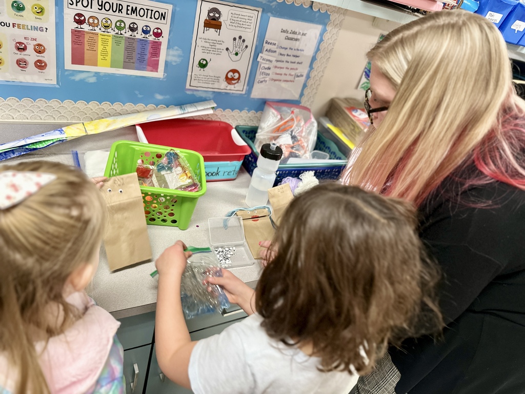 Children gather with a teacher at a counter filled with craft materials and googly eyes during a classroom activity.