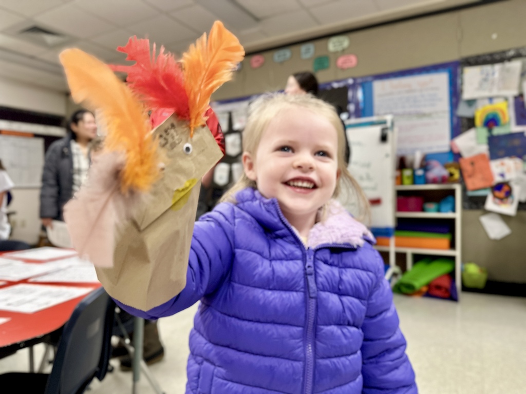 A smiling young girl in a purple coat proudly holds up a decorated paper bag puppet with colorful feathers.