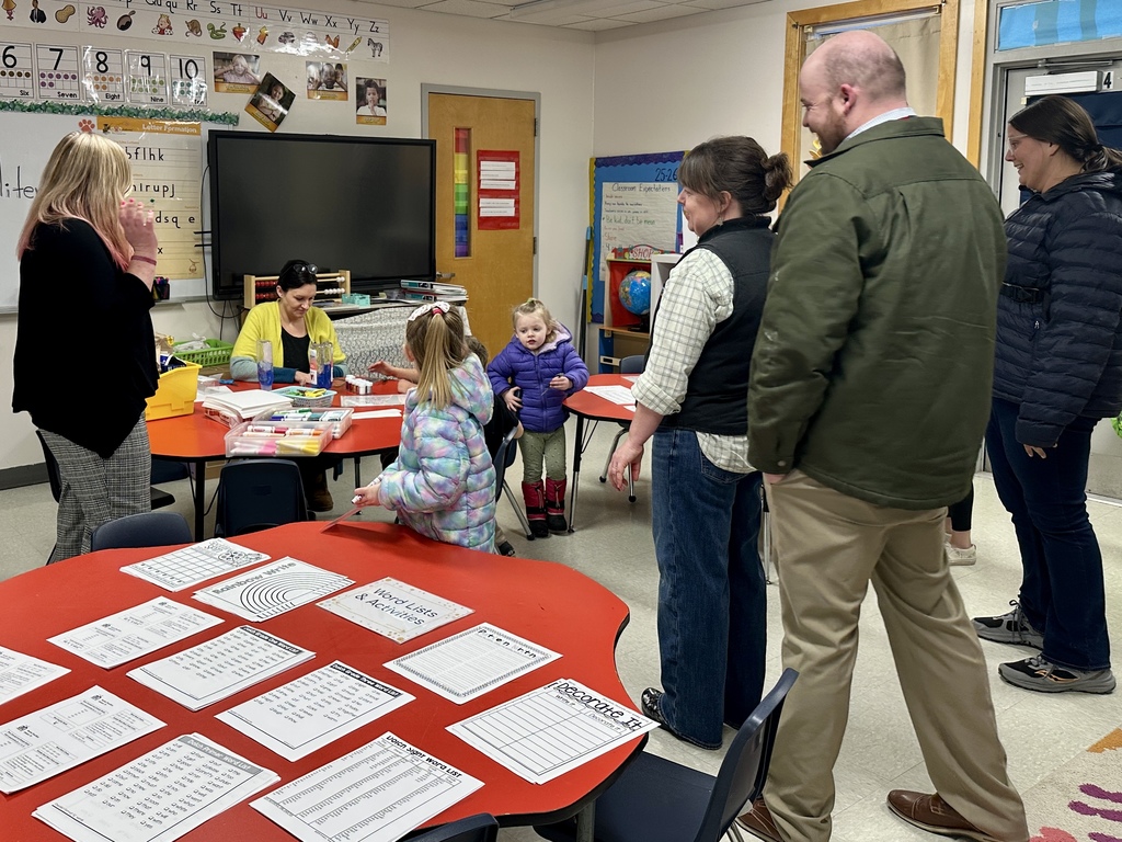 Families gather around a classroom table as a teacher works with two young girls on a sensory activity. Worksheets are laid out on nearby tables.