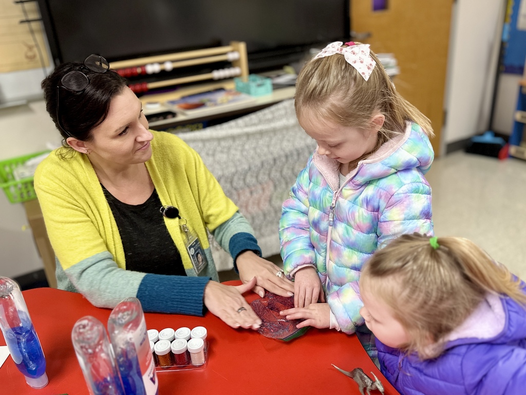 A teacher in a yellow sweater helps two young girls press their hands onto a textured sensory pad at a red table.