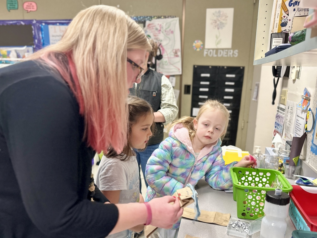A teacher with pink-tinted hair helps two girls explore craft materials at a classroom counter.