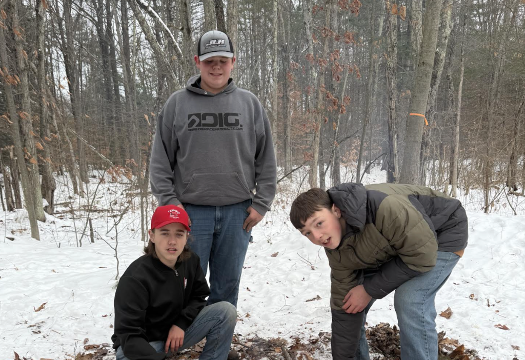 Three middle school boys outdoors in a snowy wooded area. One boy wearing a gray hoodie and baseball cap stands in the center, while another boy in a red hat kneels beside a small pile of leaves. A third boy in a green and black winter coat bends down toward the ground. Bare trees surround them in the background.