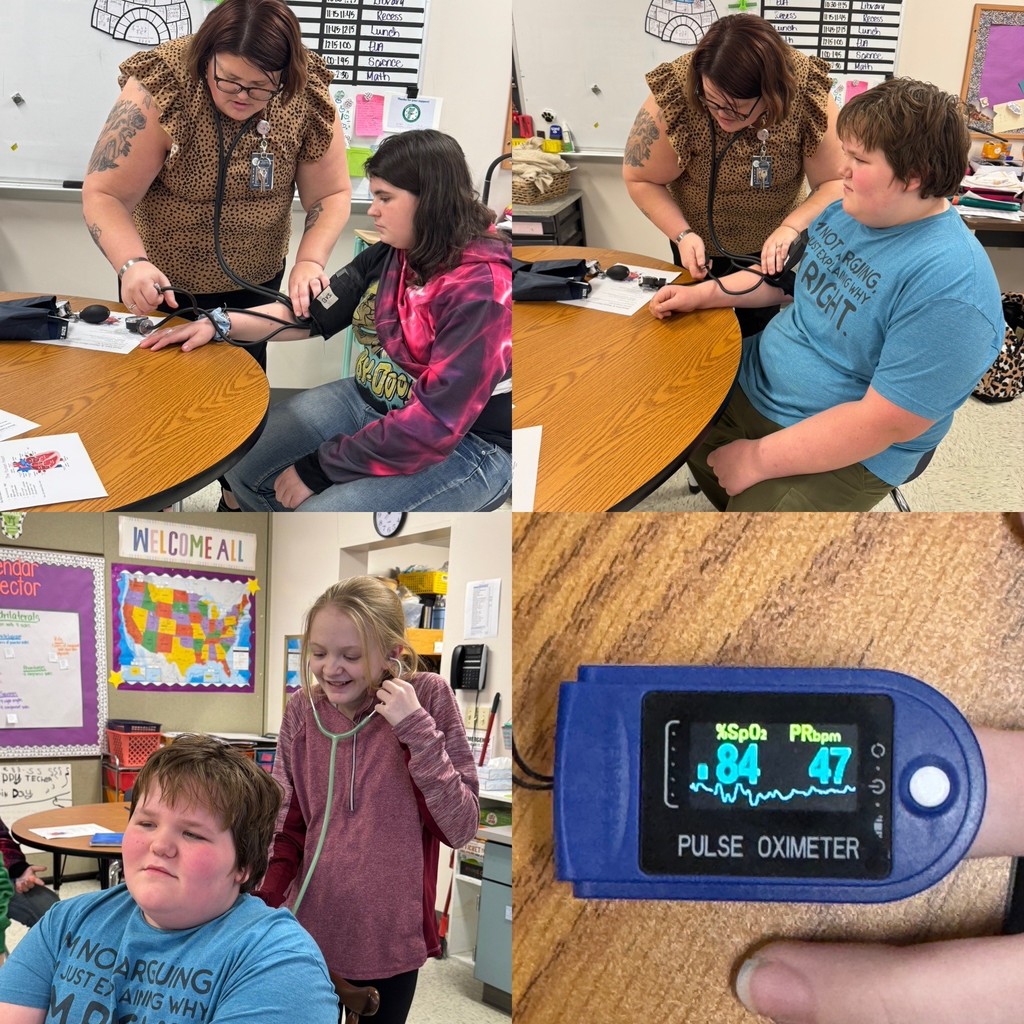 Top Left: A teacher, wearing a leopard-print top, is using a blood pressure cuff and stethoscope to take the blood pressure of a female student at a round wooden table. The student is wearing a pink and black tie-dye hoodie. Classroom posters are visible in the background. Top Right: The same teacher is taking the blood pressure of a male student at the same table. The student is wearing a blue t-shirt that says, "NOT CRYING, IM JUST IMAGINING WHY I MIGHT." Bottom Left: A female student, wearing a pink hoodie, is listening to the chest of the male student in the blue t-shirt using a stethoscope. Both are seated at a round table. A "Welcome All" poster with a map of the United States is behind them. Bottom Right: A close-up shot of a fingertip pulse oximeter displaying the results. The screen shows %SpO₂ 84 (Oxygen Saturation) and PRbpm 47 (Pulse Rate per minute). The device is blue and black.