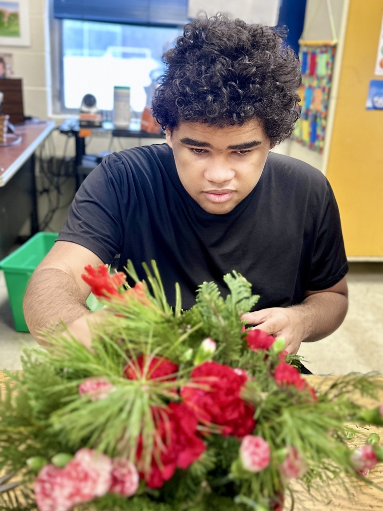 A student wearing a black t-shirt arranges a large floral display of red and pink carnations and evergreen branches. The photo focuses on the flowers in the foreground with the student working behind them.