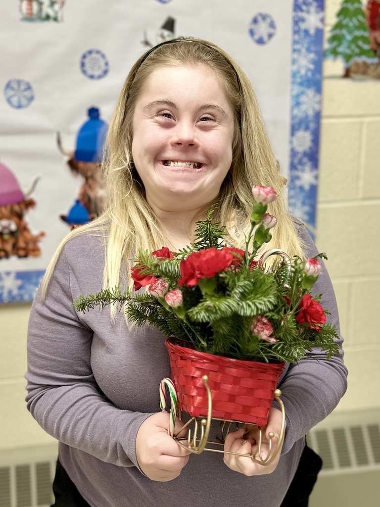 A smiling student proudly holds her finished holiday floral arrangement in a red basket sleigh. The background features winter-themed classroom decorations.