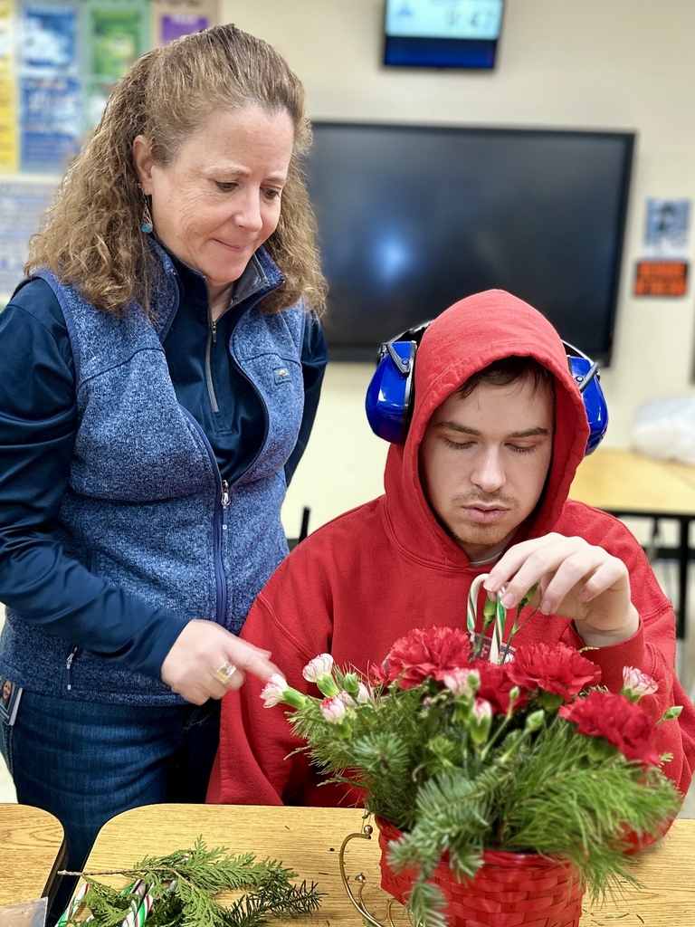 An adult wearing a blue vest smiles supportively while standing beside a student in a red hoodie with noise-canceling headphones. The student carefully places flowers into a holiday-themed arrangement.