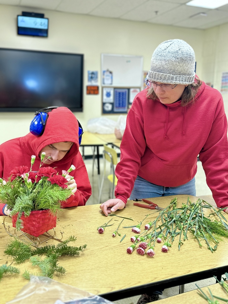 A student in a red hoodie with blue noise-canceling headphones arranges flowers in a red basket while an adult in a gray knit hat stands nearby offering support. Carnations and greenery are spread out on the table.