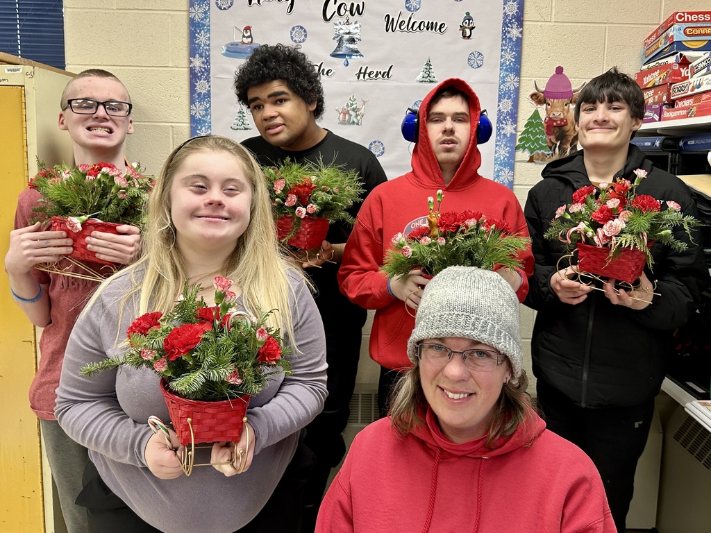 A group of students and a staff member pose together holding their completed floral arrangements. Each student holds a red sleigh-shaped basket filled with bright holiday flowers and greenery. Everyone smiles proudly at the camera.