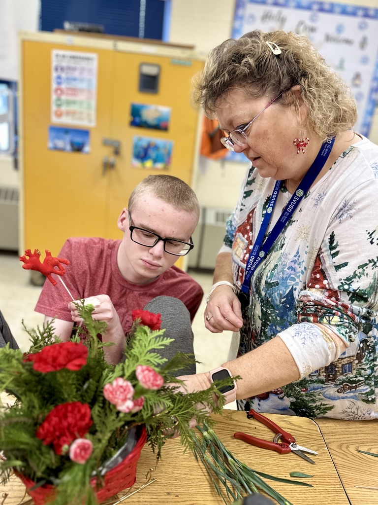A student wearing glasses and a red shirt focuses intently while creating a floral arrangement. An adult standing beside him gives guidance. The red and pink carnations and greenery fill a red basket on the table.