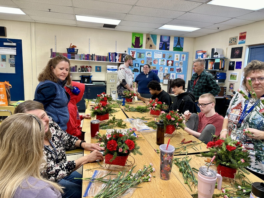 A wide classroom view shows several students and staff working together at long tables to assemble festive floral arrangements. Red baskets filled with carnations and greenery are spread across the tables.