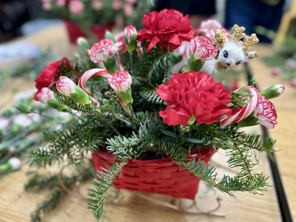 A close-up of a completed floral arrangement featuring red carnations, pink-tipped carnations, evergreen branches, and a small decorative figurine tucked among the flowers inside a red basket sleigh.