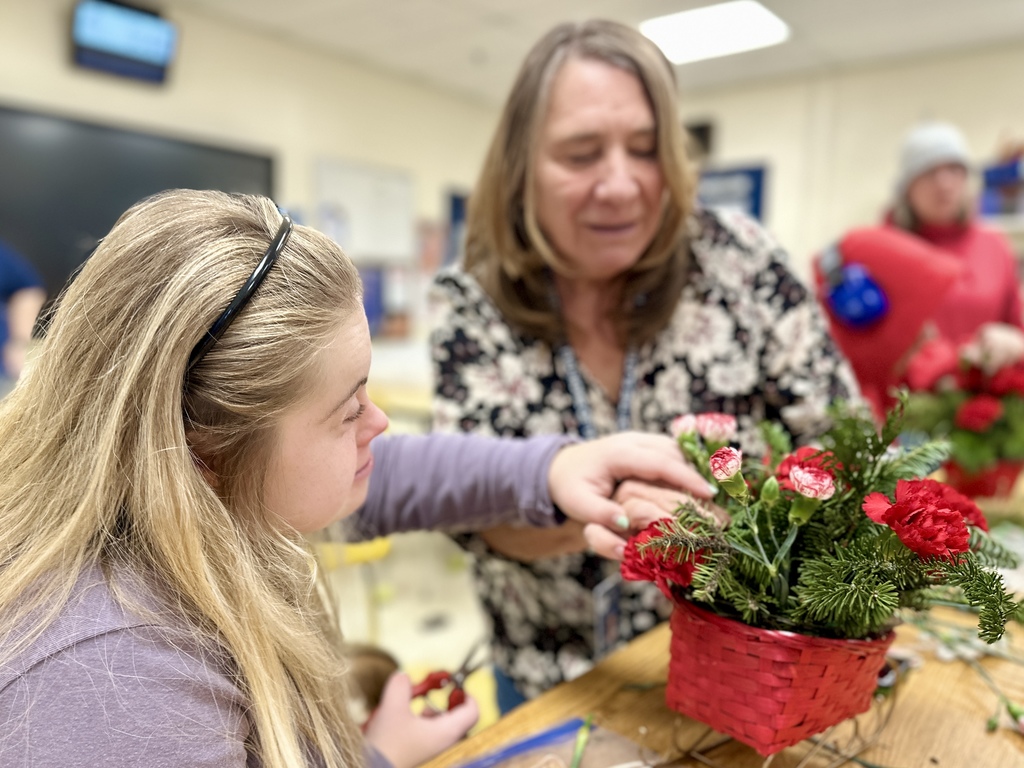 A student with long blonde hair works on a holiday floral arrangement alongside an adult. The adult gently guides the student’s hand as they arrange red and pink carnations and evergreens in a red basket. The background shows a classroom setting.