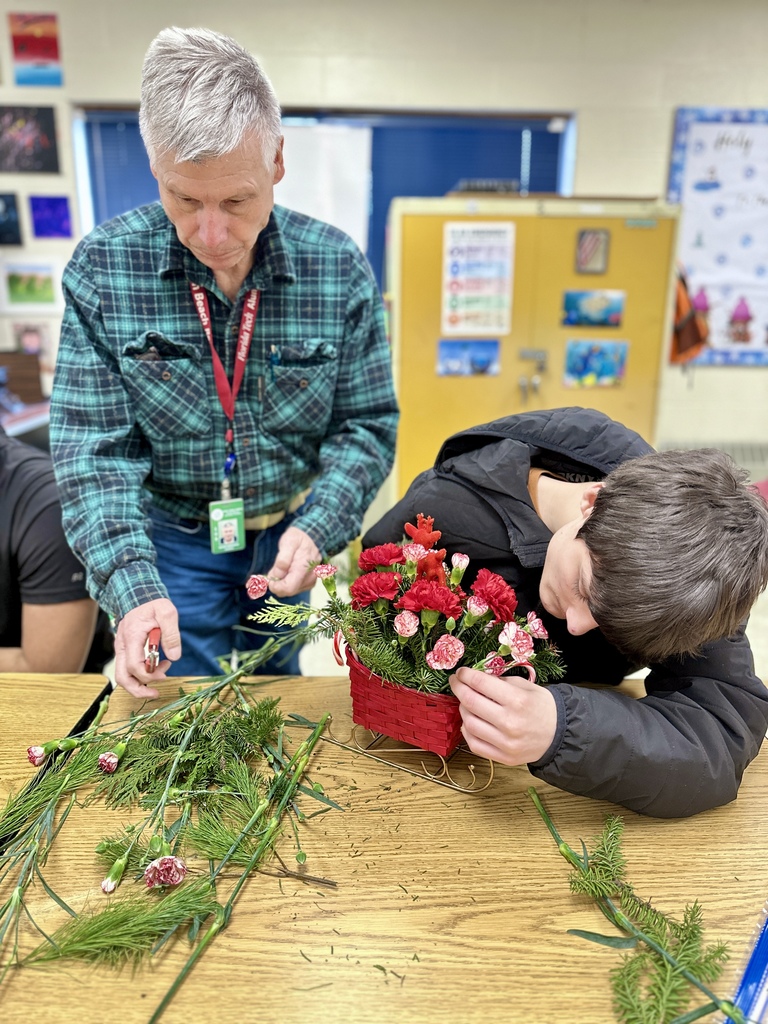 An adult in a green flannel shirt helps a student adjust flowers in a red basket arrangement. Evergreen branches and flower stems are scattered across the table as they work.