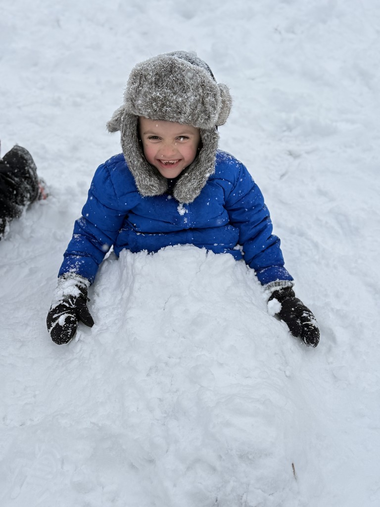 A young boy in a bright blue winter coat and furry gray hat grins while sitting in deep snow, his black gloves resting on top.