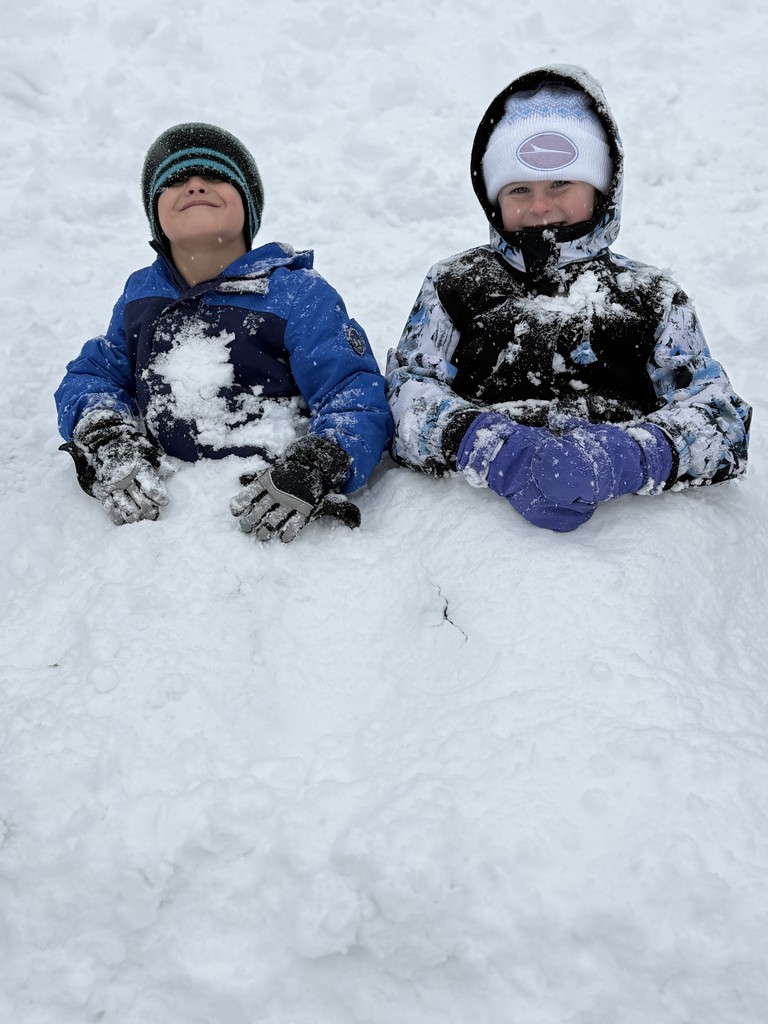 Two children lie back in the snow, buried up to their waists. The child on the left wears a blue coat and striped hat with a playful grin, while the child on the right wears a patterned winter coat and white Slater beanie, smiling warmly.