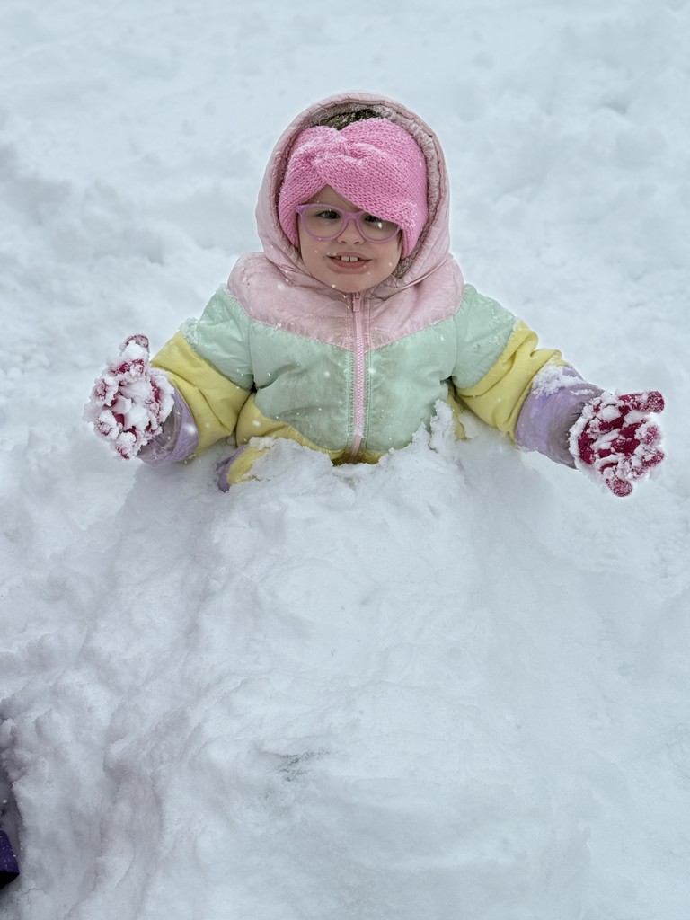 A young girl wearing a pastel rainbow puffer jacket, pink knit headband, and pink glasses sits in deep snow, smiling with her gloved hands covered in snow.