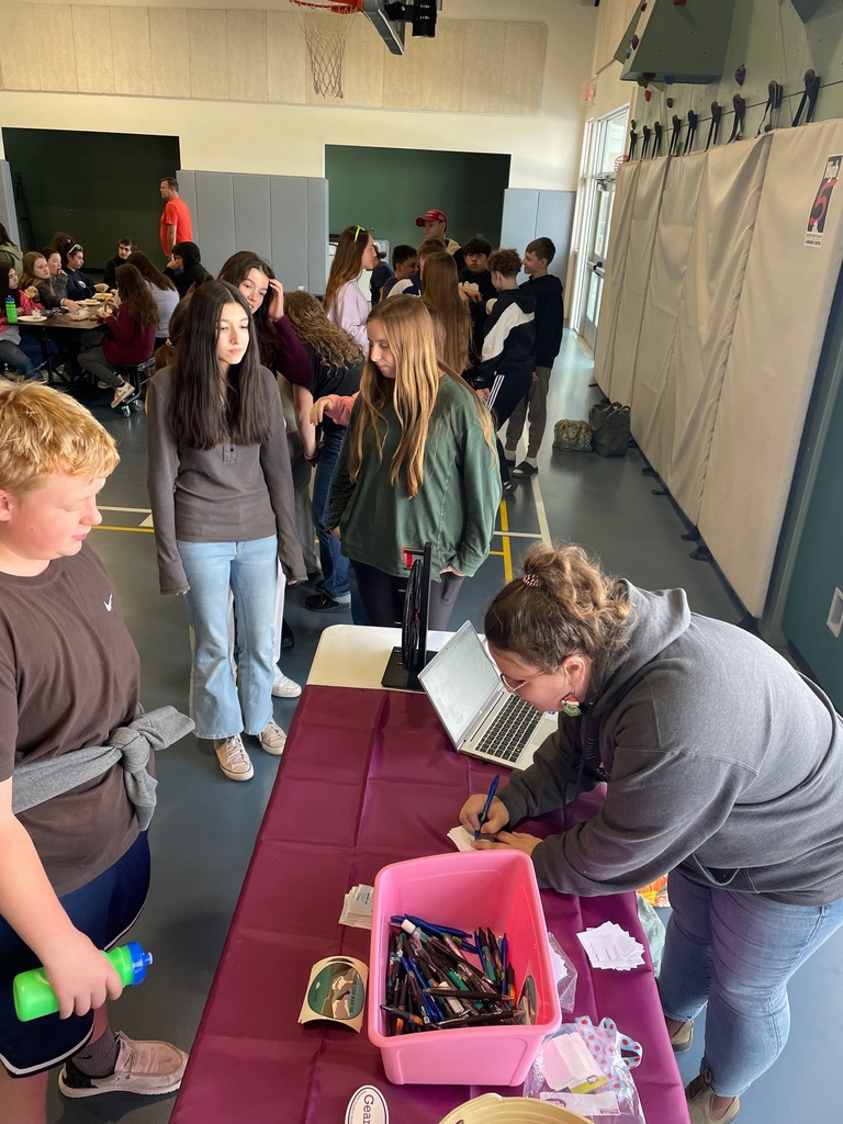 Students stand in line at a table covered with a purple cloth in a school gym, while a staff member leans over a laptop to help them sign in. A pink bin filled with pens sits on the table.